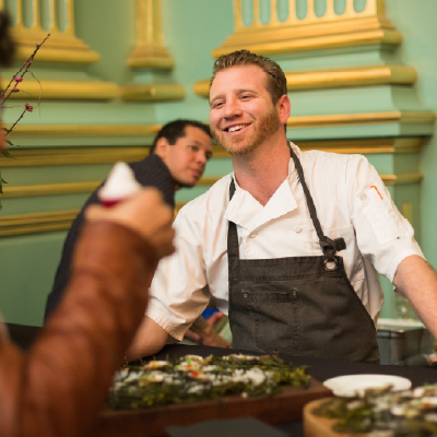 A cheerful chef in a white uniform interacting with guests while presenting gourmet dishes.